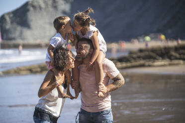 Sibling kissing while sitting on parents shoulder at beach - SNF01152