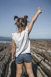 Girl gesturing peace sign while standing on flysch against blue sky during sunny day - SNF01154