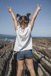 Girl with arms raised gesturing peace sign while standing on flysch against sky - SNF01155