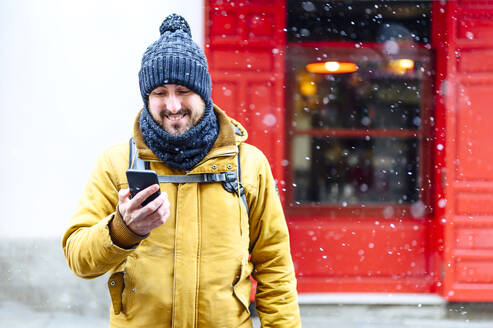 Lächelnder mittelgroßer erwachsener Mann, der auf sein Mobiltelefon schaut, während er im Winter vor einem Geschäft steht - PGF00407