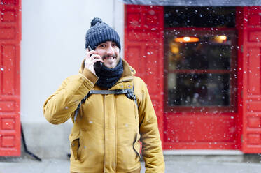 Smiling man talking on mobile phone while standing against store during winter - PGF00408