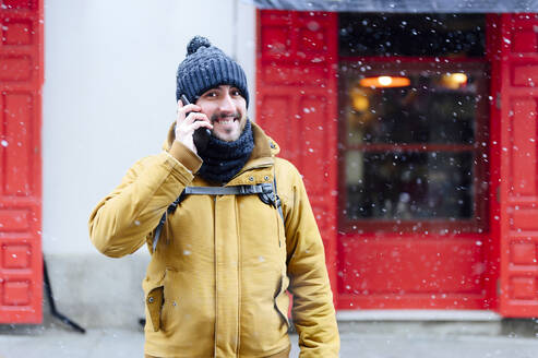 Lächelnder Mann, der mit seinem Handy telefoniert, während er im Winter vor einem Geschäft steht - PGF00408