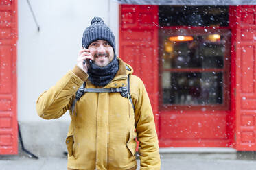 Smiling man in warm clothing talking on smart phone while snowing during winter - PGF00409