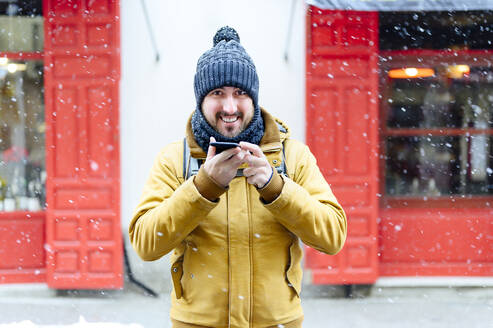 Glücklicher Mann, der sein Mobiltelefon gegen ein Geschäft hält, während es im Winter schneit - PGF00410