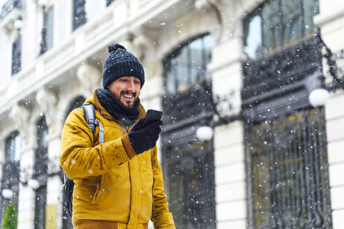 Smiling man looking at mobile phone by building while snowing during winter - PGF00414