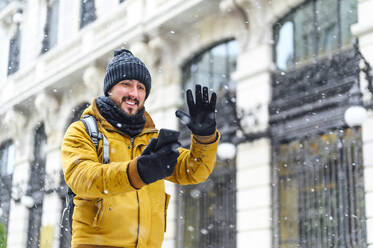 Smiling man talking on video call through mobile phone by building while snowing during winter - PGF00415