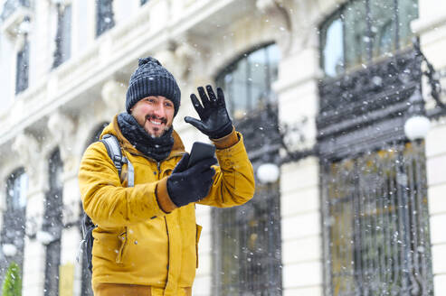 Lächelnder Mann winkt mit der Hand zum Mobiltelefon, während er einen Videoanruf bei einem Gebäude im Winter führt - PGF00416