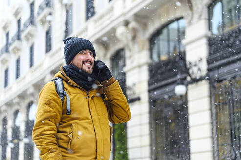 Lächelnder Mann in warmer Kleidung, der im Winter mit seinem Smartphone an einem Gebäude telefoniert und dabei wegschaut - PGF00417