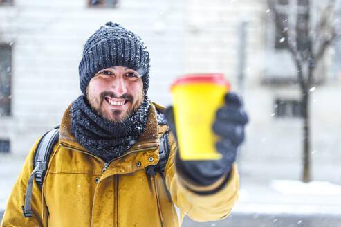 Glücklicher Mann zeigt Kaffeetasse in die Kamera, während es im Winter schneit - PGF00418