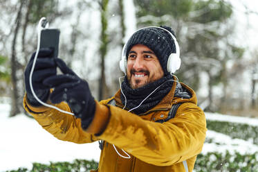 Happy man with headphones listening music while taking selfie through mobile phone during winter - PGF00419