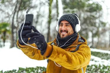 Mid adult man with headphones listening music while taking selfie through smart phone on snow covered land during winter - PGF00420