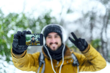 Mann gestikuliert bei der Aufnahme eines Selfies mit dem Handy im Winter - PGF00421