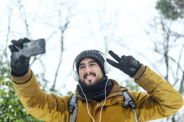Smiling man in warm clothing listening music while taking selfie through mobile phone against sky - PGF00422