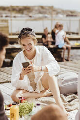 Smiling woman using smart phone sitting by friend on jetty during picnic - MASF21768