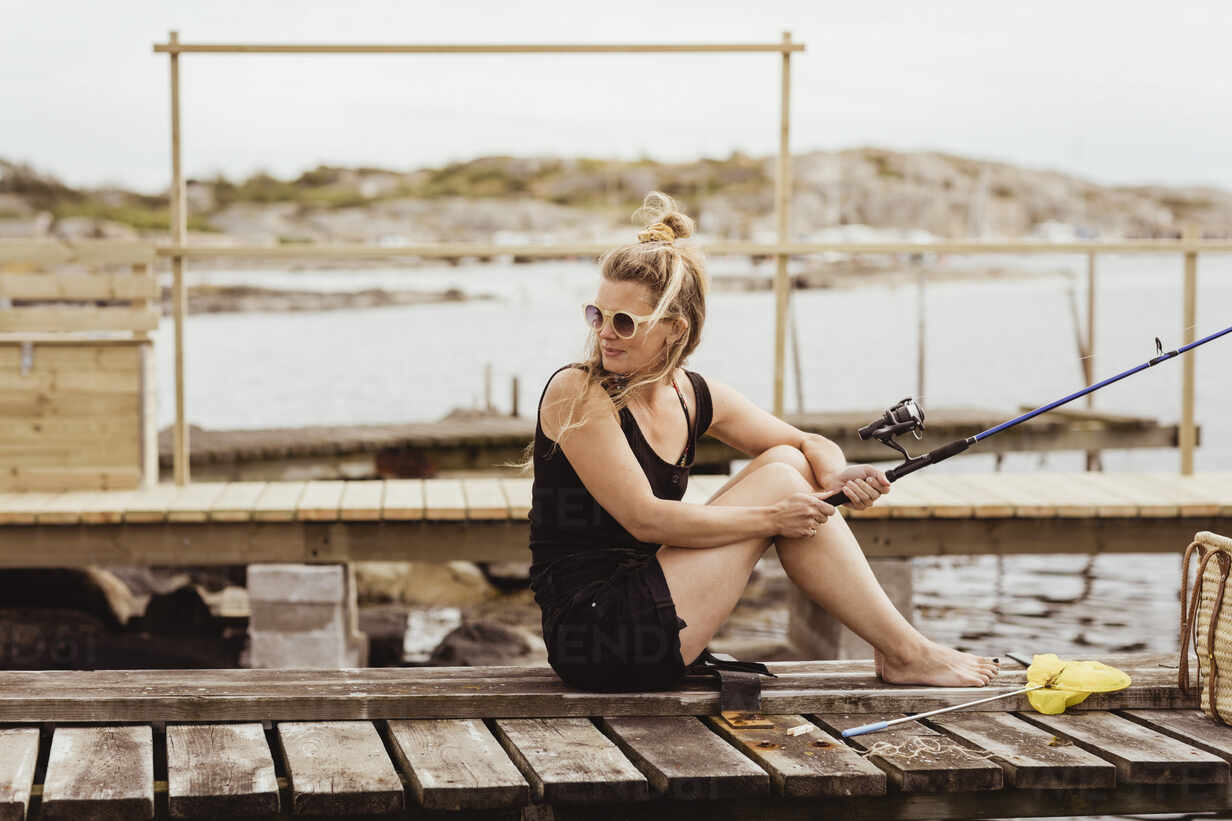 Mature woman with fishing rod looking over shoulder while sitting on jetty  at harbor stock photo