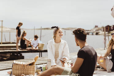 Smiling friends talking while sitting over jetty on sunny day during picnic - MASF21776