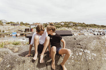 Happy siblings sitting on rock against sky during summer vacation - MASF21788