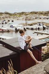 Smiling boy with fishing rod sitting on rooftop at harbor during vacation - MASF21797