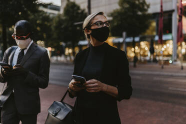 Unternehmerin mit Smartphone schaut weg, während sie bei COVID-19 neben einem männlichen Kollegen steht - MASF21804