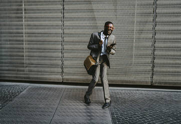 Businessman laughing while standing with smart phone on walkway against metal grate - MASF21826