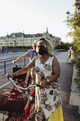 Senior man and woman wheeling bicycle on bridge during pandemic - MASF21863