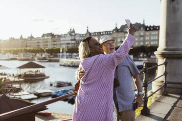Lächelnde ältere Paar unter selfie während stehend auf Brücke gegen See in der Stadt - MASF21867