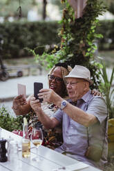 Cheerful senior couple taking selfie on smart phone while sitting in sidewalk cafe - MASF21898