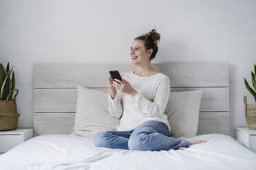 Lächelnde Frau, die ein Mobiltelefon benutzt, während sie zu Hause auf dem Bett sitzt - EBBF02415
