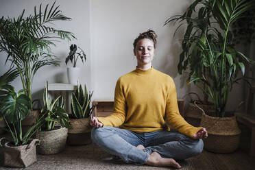 Young woman meditating while sitting on floor by plant at home - EBBF02424
