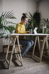 Lächelnde Frau, die an einem digitalen Tablet arbeitet, während sie an einem Tisch im Heimbüro sitzt - EBBF02428