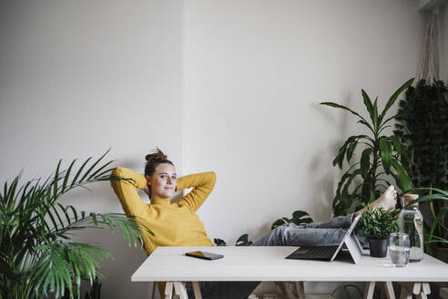 Smiling woman with hands behind head resting while sitting at home office - EBBF02432