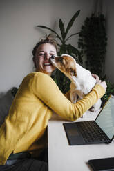 Smiling woman playing with dog while sitting at home office - EBBF02435