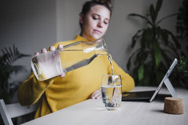 Frau gießt Wasser in ein Glas, während sie mit einem digitalen Tablet im Büro zu Hause sitzt - EBBF02441