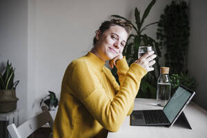 Smiling woman drinking water while using digital tablet sitting at home office - EBBF02442