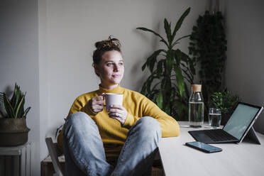Junge Frau trinkt Kaffee, während sie am Tisch im Heimbüro sitzt - EBBF02445