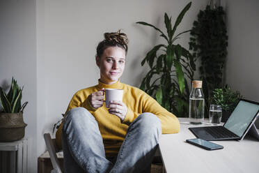 Lächelnde Frau trinkt Kaffee, während sie zu Hause im Büro sitzt - EBBF02446