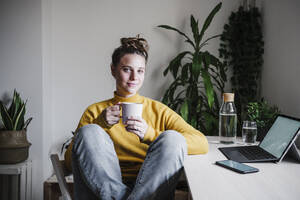 Smiling woman drinking coffee while sitting at home office - EBBF02446