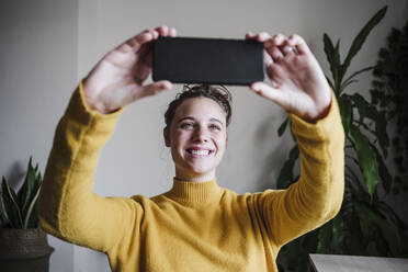 Junge Frau lächelt, während sie ein Selfie mit ihrem Mobiltelefon macht und zu Hause sitzt - EBBF02447