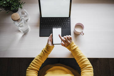 Frau, die ein Mobiltelefon benutzt, während sie mit einem digitalen Tablet auf dem Tisch im Büro zu Hause sitzt - EBBF02448