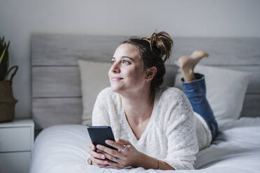 Lächelnde Frau, die ein Mobiltelefon benutzt, während sie auf dem Bett liegt - EBBF02450