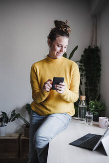 Junge Frau, die ein Mobiltelefon benutzt, während sie mit einem digitalen Tablet auf einem Tisch zu Hause sitzt - EBBF02451