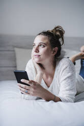 Nachdenkliche Frau mit Hand am Kinn, die ein Mobiltelefon benutzt, während sie zu Hause auf dem Bett liegt - EBBF02456