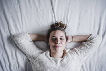 Smiling woman with hands behind head resting while lying on bed at home - EBBF02464