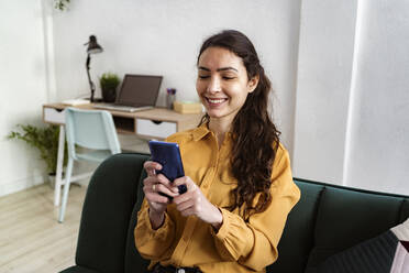 Young woman smiling while using mobile phone sitting on sofa at home - GIOF10998