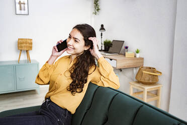 Woman talking on mobile phone while sitting on sofa at home - GIOF11000