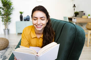 Young woman reading book while lying on sofa at home - GIOF11006