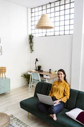 Woman using laptop while sitting on sofa at home - GIOF11009