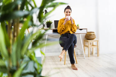 Young woman smiling while drinking coffee sitting on stool at home - GIOF11016