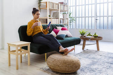 Frau mit Kaffeetasse und Mobiltelefon, während sie zu Hause auf dem Sofa sitzt - GIOF11021