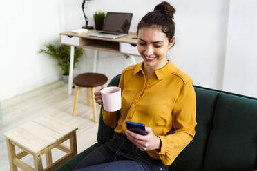 Lächelnde junge Frau, die einen Kaffee trinkt und dabei ein Mobiltelefon benutzt, während sie zu Hause sitzt - GIOF11022